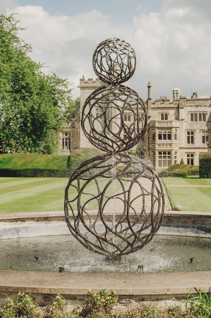 A modern spherical metal sculpture stands in a fountain with water spraying out, creating a picturesque scene perfect for a Farleigh House Wedding Photographer, set against the backdrop of a historic building and manicured lawn.