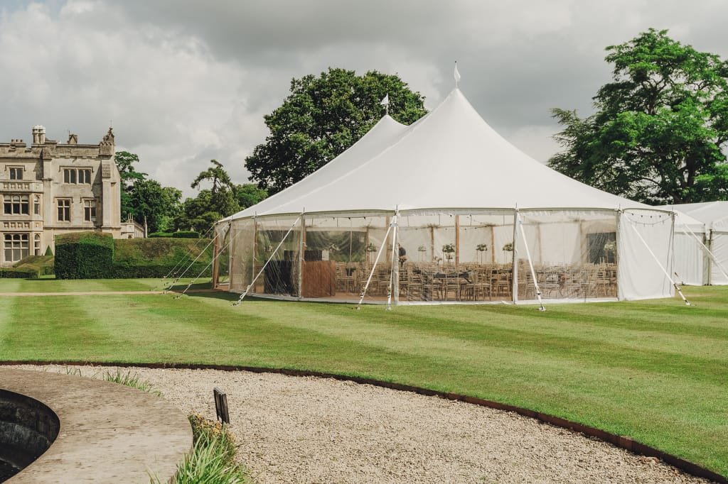 A white event tent is pitched on a well-manicured lawn beside the historic Farleigh House. The interior of the tent is visible, showing tables and chairs set up for an event, perfect for capturing by a wedding photographer. The sky is cloudy.