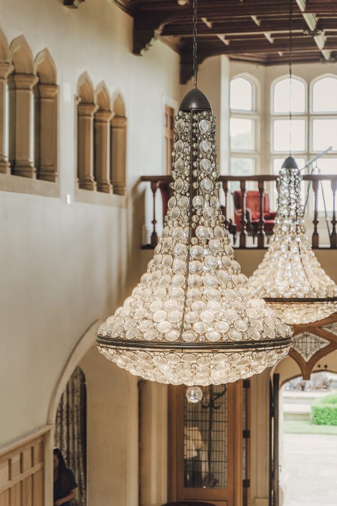 A grand, multi-tiered crystal chandelier hangs in a high-ceilinged, elegantly decorated room with arched windows and doorways, perfect for a Farleigh House wedding photographer to capture stunning moments.