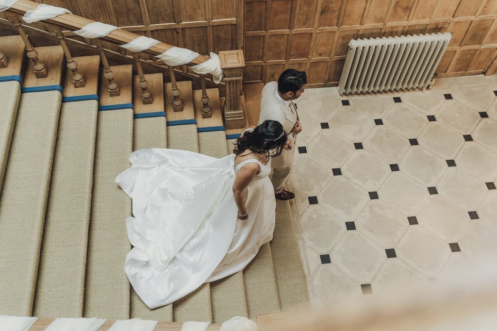 Bride and groom walking down a grand wooden staircase with a beige carpet, the bride wearing a flowing white wedding gown. Captured beautifully by Farleigh House Wedding Photographer.