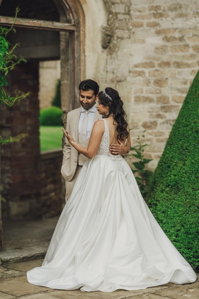 A bride and groom stand together outside, the bride in a white gown and the groom in a beige suit, with a stone building and greenery in the background, captured beautifully by their Farleigh House Wedding Photographer.