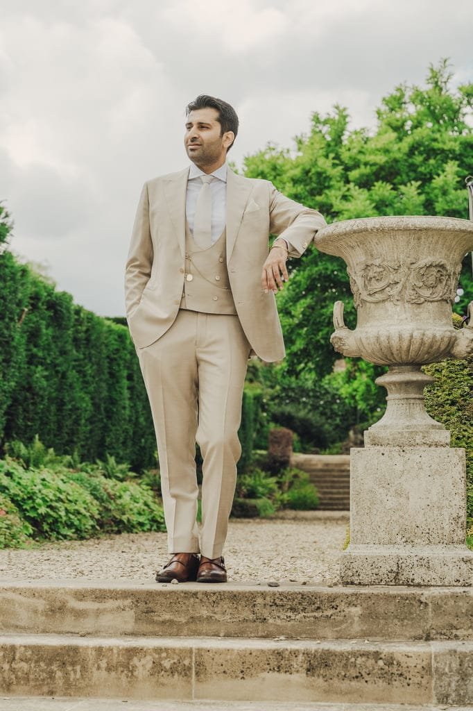 A man in a beige suit and brown shoes stands on a stone path, leaning on a large decorative urn, with greenery and stone steps in the background—the perfect scene for a Farleigh House Wedding Photographer.