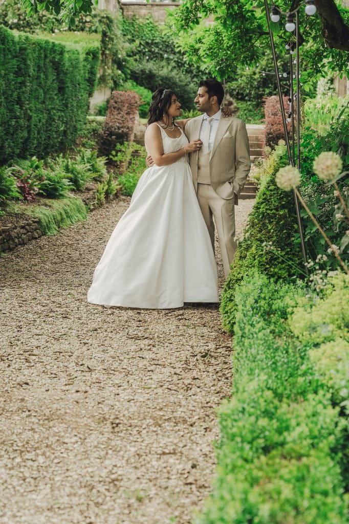 A couple in wedding attire stands closely together, looking at each other, on a gravel pathway surrounded by lush green gardens, captured beautifully by a Farleigh House Wedding Photographer.
