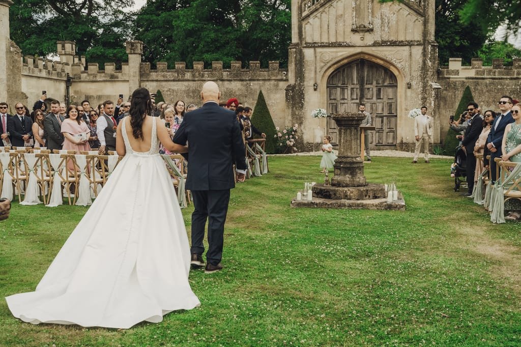 A bride in a white dress and a man in a suit walk down an outdoor aisle toward a group of seated guests, with the historical Farleigh House as their backdrop, captured beautifully by their wedding photographer.