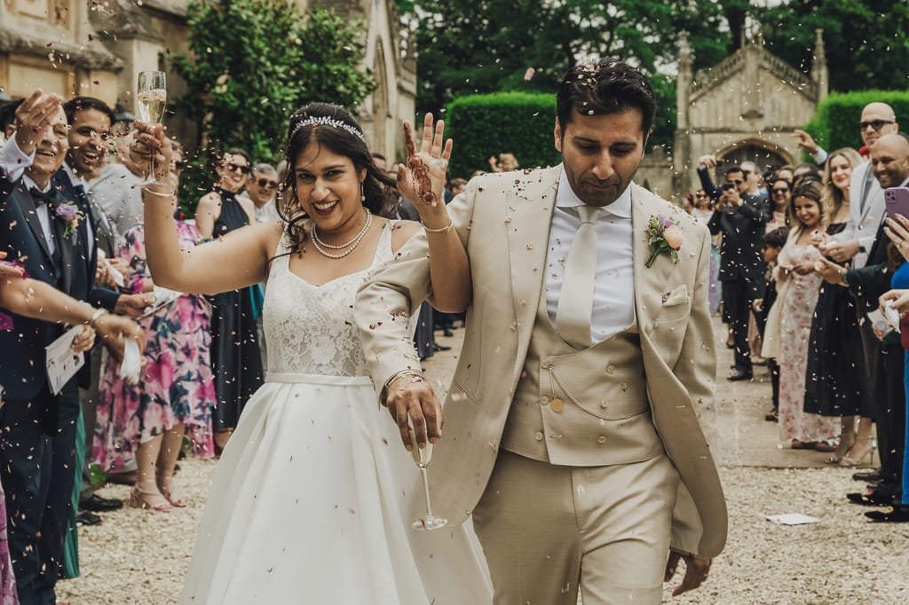 A bride and groom walk down an outdoor path at Farleigh House as guests shower them with confetti. The bride is smiling and holding a champagne glass, while the groom in a tan suit walks beside her, both perfectly captured by their wedding photographer.