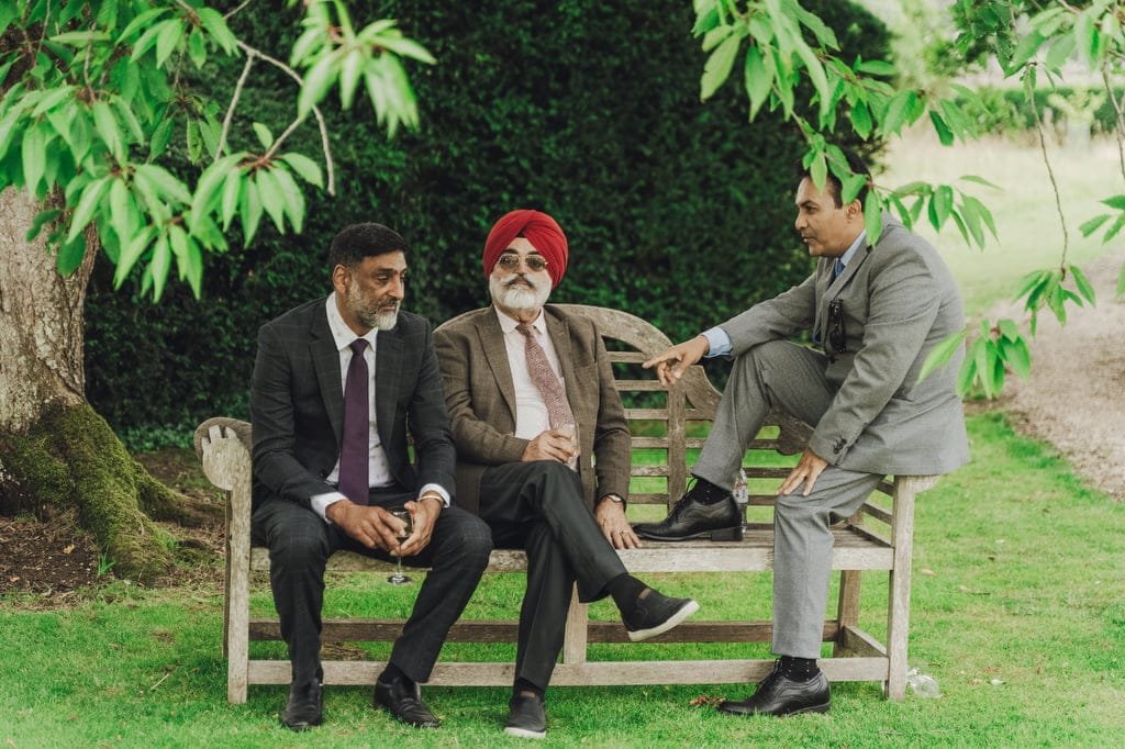 Three men in formal attire sit and converse on a wooden bench under green foliage, with one man wearing a red turban. The scene captures the charm that a Farleigh House Wedding Photographer would beautifully document.