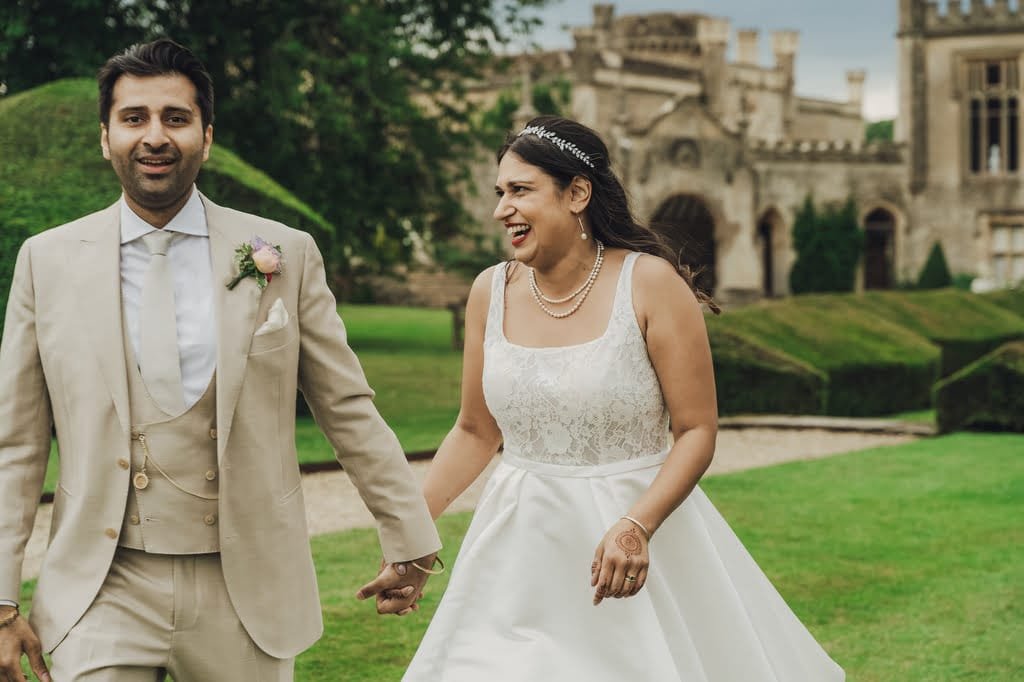 A couple in wedding attire holds hands and smiles while walking in an outdoor garden with Farleigh House in the background, captured beautifully by their wedding photographer.