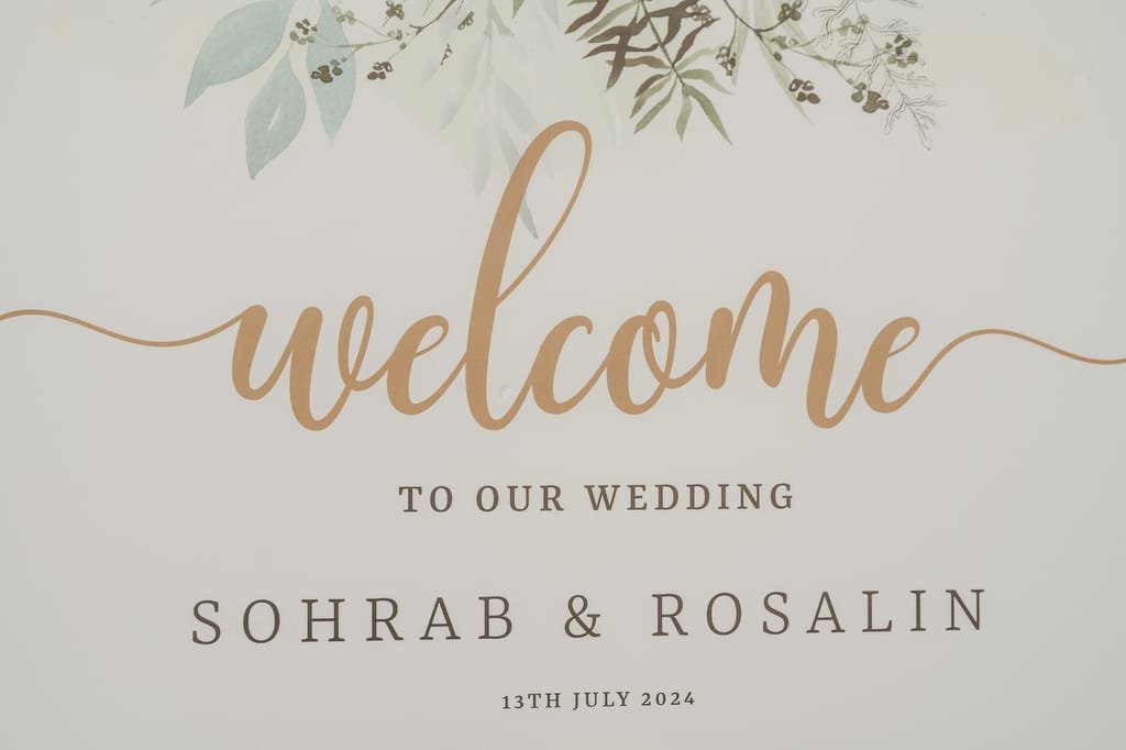 A wedding welcome sign reads, "Welcome to our wedding," followed by the names "Sohrab & Rosalin" and the date "13th July 2024," with floral illustrations at the top. Captured beautifully by Farleigh House Wedding Photographer.