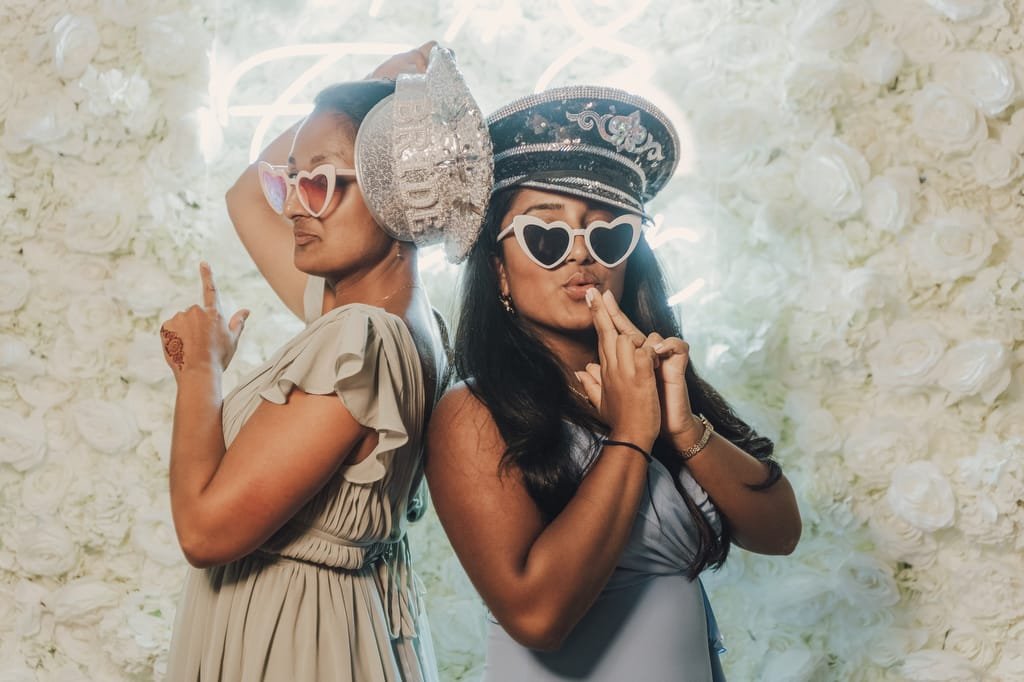 Two people posing in front of a floral background, wearing heart-shaped sunglasses and holding playful props, one with a decorative hat and the other with a silver hat—a charming moment captured by Farleigh House Wedding Photographer.