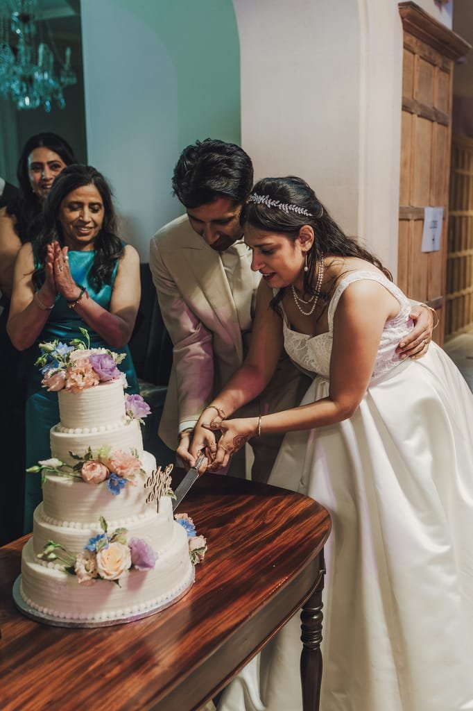 A bride and groom cut a three-tiered wedding cake adorned with pastel-colored flowers, while onlookers in formal attire smile and clap. Captured beautifully by the Farleigh House Wedding Photographer, every magical moment is immortalized.
