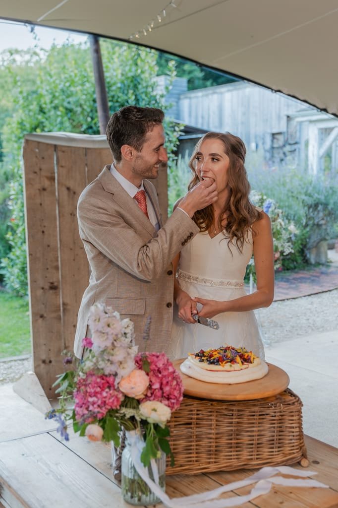 A groom in a light beige suit feeds a piece of wedding cake to a bride in a white dress as they stand next to a decorated cake on a wicker stand at PYTHOUSE WEDDINGS. A bouquet of flowers is in the foreground.