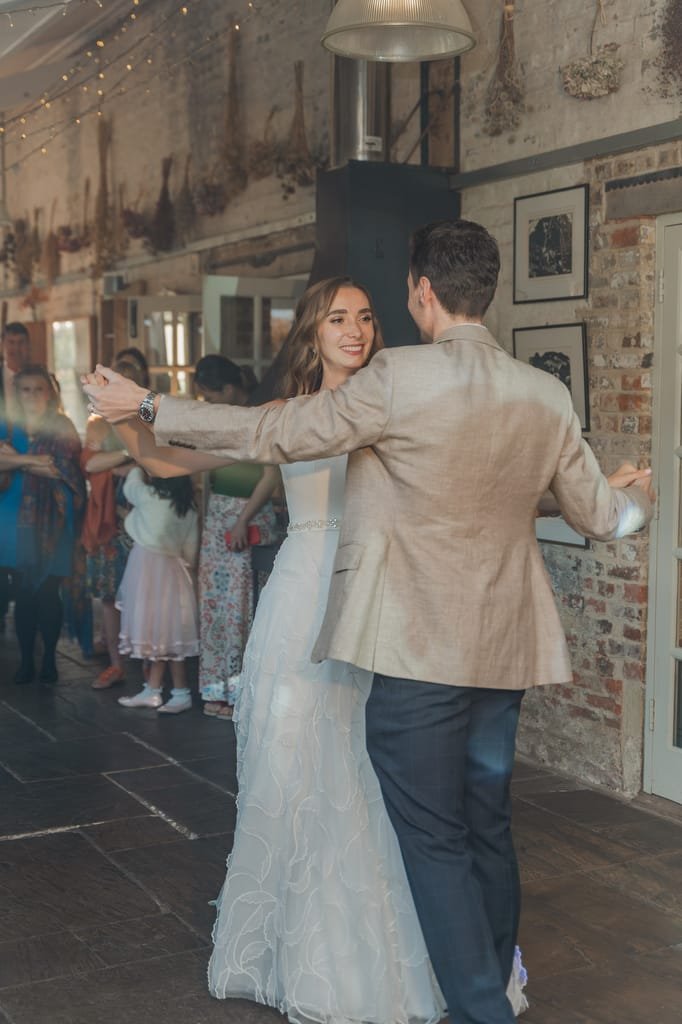 A couple dances in a warmly lit room with rustic decor at Pythouse Weddings, while a small crowd watches. The woman wears a white gown, and the man wears a beige jacket and dark pants.