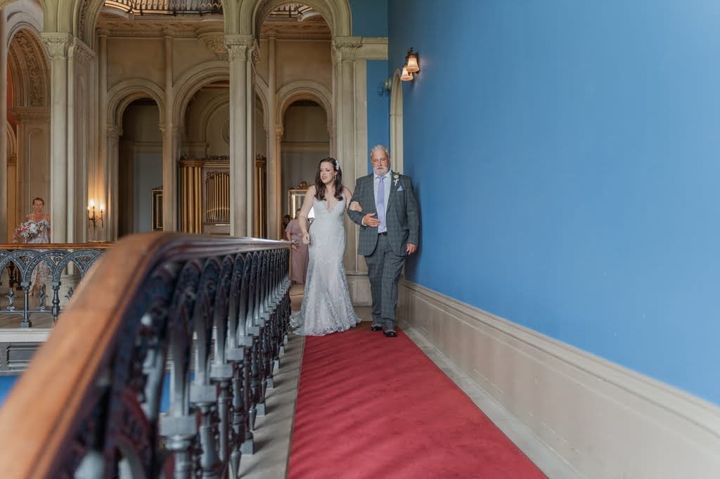 A bride in a white gown walks down a carpeted hallway accompanied by an older man in a suit, captured beautifully by the Grittleton House Photographer, within an ornate building with blue walls and archways.