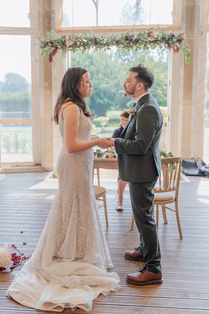 A bride and groom face each other, holding hands in a brightly lit room with large windows and floral decor at Grittleton House. The bride is in a lace gown, the groom in a dark suit. A photographer is subtly seated behind them, capturing their special moment.