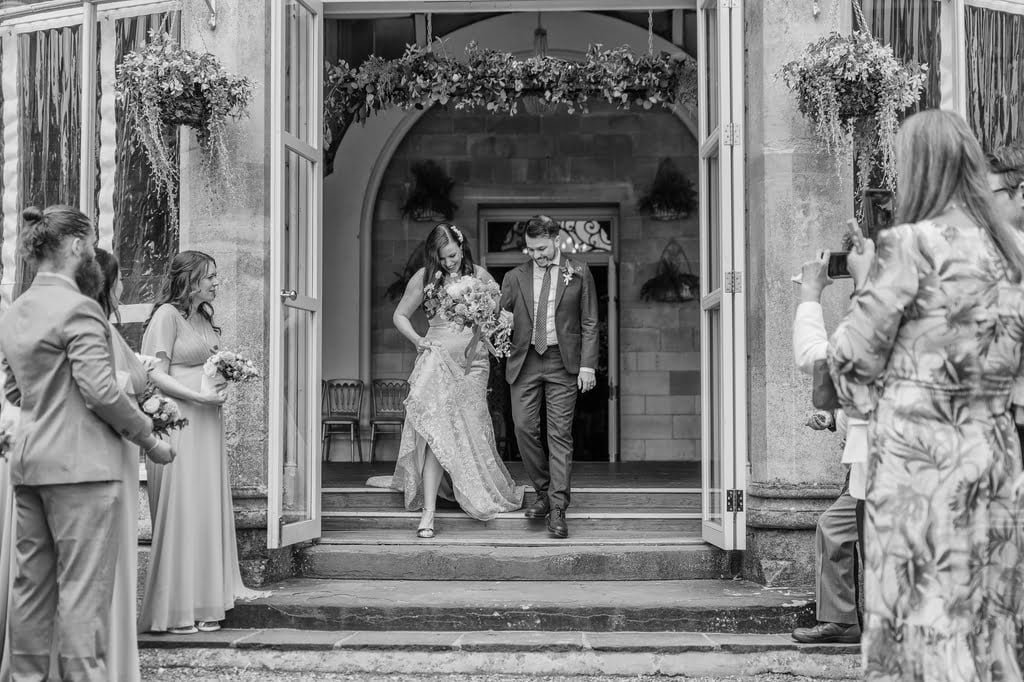 Black and white photo of a newlywed couple walking down steps at Grittleton House, surrounded by wedding guests taking pictures and wearing formal attire.