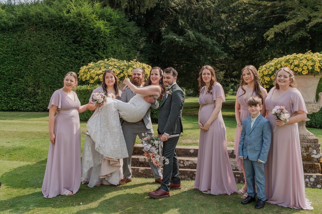 A wedding party poses outdoors on a lawn at Grittleton House, with the groom lifting the bride. Bridesmaids in lavender dresses and groomsmen in suits stand around them, along with a young boy wearing a suit, all captured beautifully by the Grittleton House Photographer.