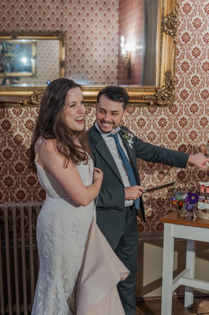 Bride and groom smiling and cutting a wedding cake with a knife, standing in front of patterned wallpaper and a large, ornate mirror, captured beautifully by our Grittleton House photographer.