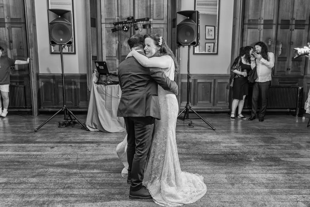 A bride and groom embrace on the dance floor during their wedding reception in a room with wooden paneling. A DJ is behind them, and other guests are socializing in the background, captured beautifully by the Grittleton House Photographer.
