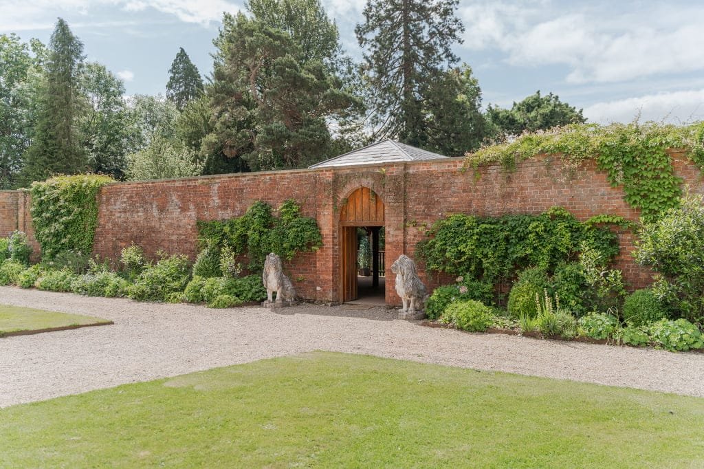 A brick wall with an arched wooden door is covered in green ivy at Elmhay Park. Two stone lion statues stand guard on either side of the entrance. The scene includes a gravel path and a grassy area in the foreground, perfect for any weddings photographer to capture timeless moments.