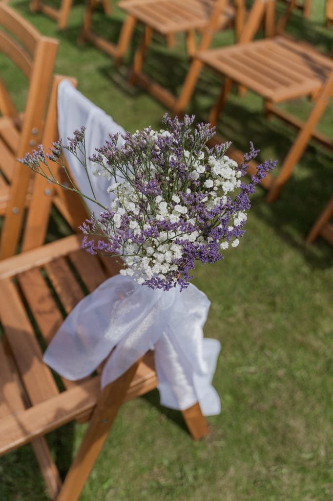 A bouquet of white and purple flowers tied to a wooden chair with a white ribbon, set outdoors on a grassy lawn at Elmhay Park — the perfect shot for any weddings photographer.