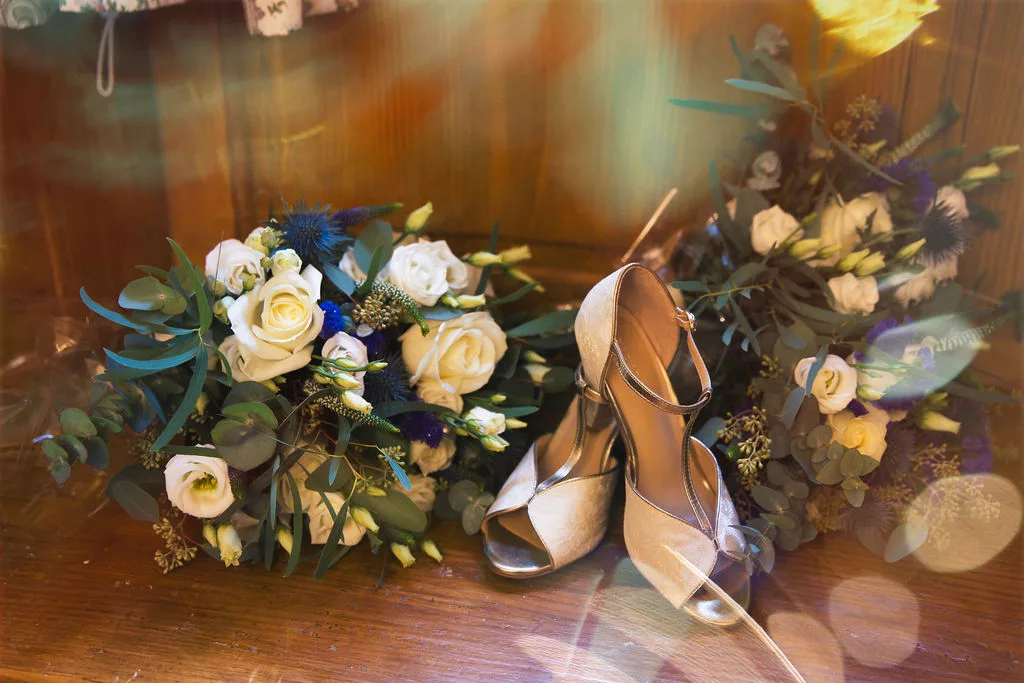 A bride's wedding shoes and bouquet on a wooden table captured by a Wedding Photographer in Bath.