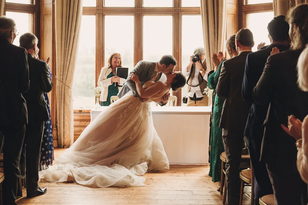 A bride and groom kiss passionately by a window in a warmly lit room during their Orchardleigh House wedding as guests applaud, with a photographer capturing the moment.