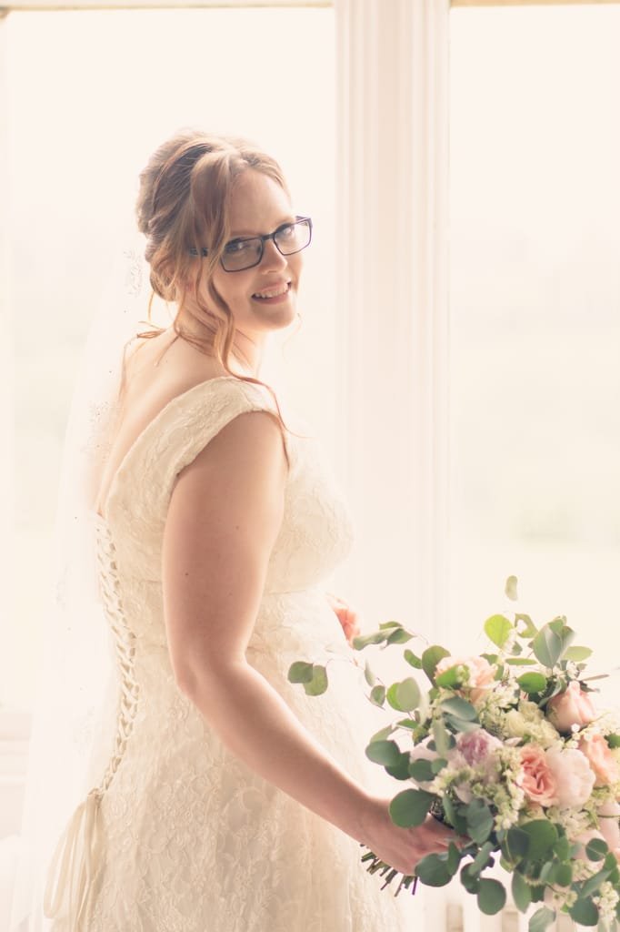 A bride in a lace dress and glasses smiling by a window, holding a bouquet of pink and green flowers, captured beautifully by her wedding photographer at Orchardleigh.