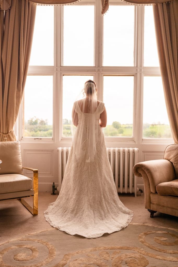 A bride in a long white gown and veil stands facing a large window overlooking a green landscape, captured by the wedding photographer at Orchardleigh, in a room with elegant decor.