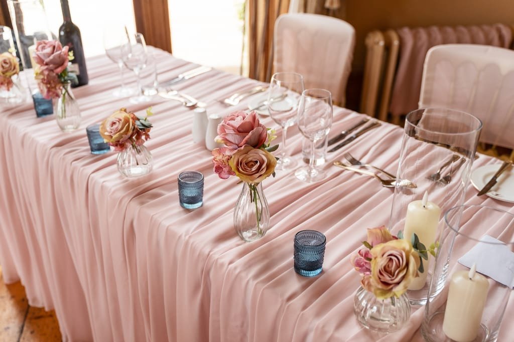 Elegant wedding dining table setup with pink tablecloth, floral centerpieces, candles, and wine glasses in a well-lit room.