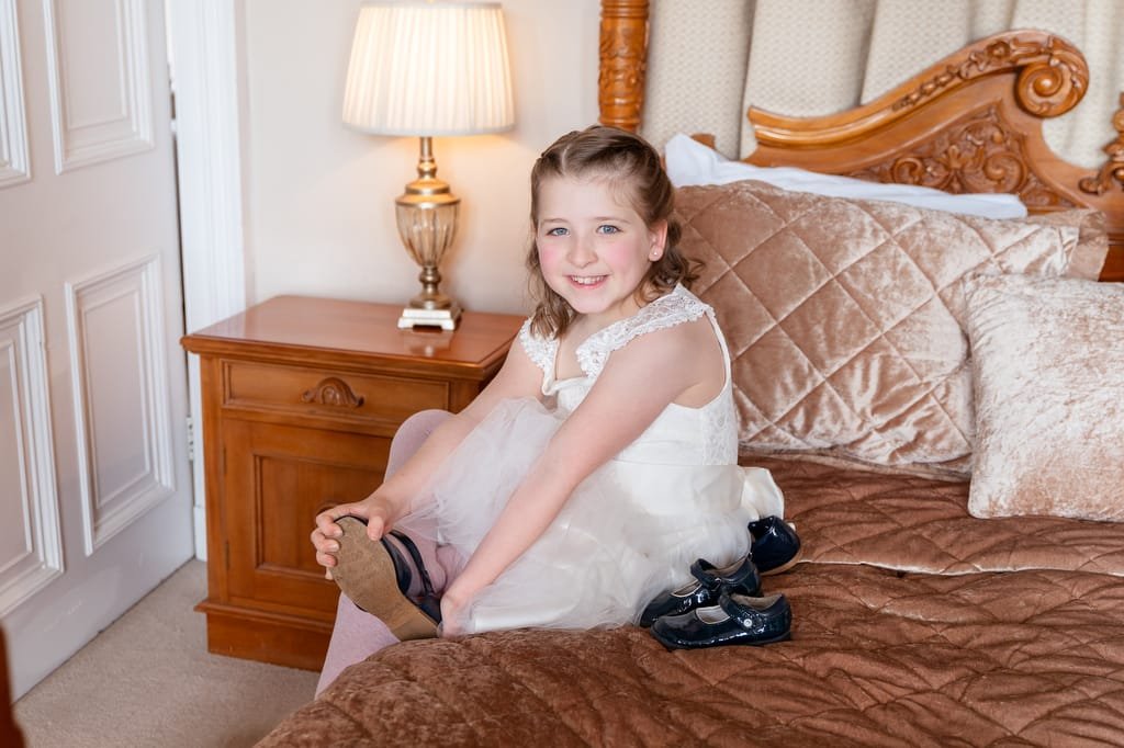 A young girl in a white dress smiling while sitting on a bed, putting on her shoes in a well-furnished room, captured by a wedding photographer at Orchardleigh.