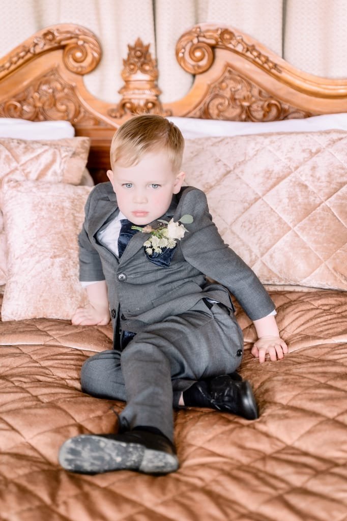 A young boy in a gray suit and black shoes sits on an ornate bed with beige bedding, looking directly at the camera, captured by a wedding photographer at Orchardleigh.