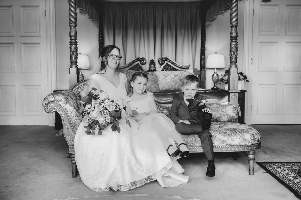 A black and white photo by Orchardleigh's wedding photographer of a bride sitting on an ornate sofa, holding a bouquet, with a young girl and a boy seated beside her in a formal room.