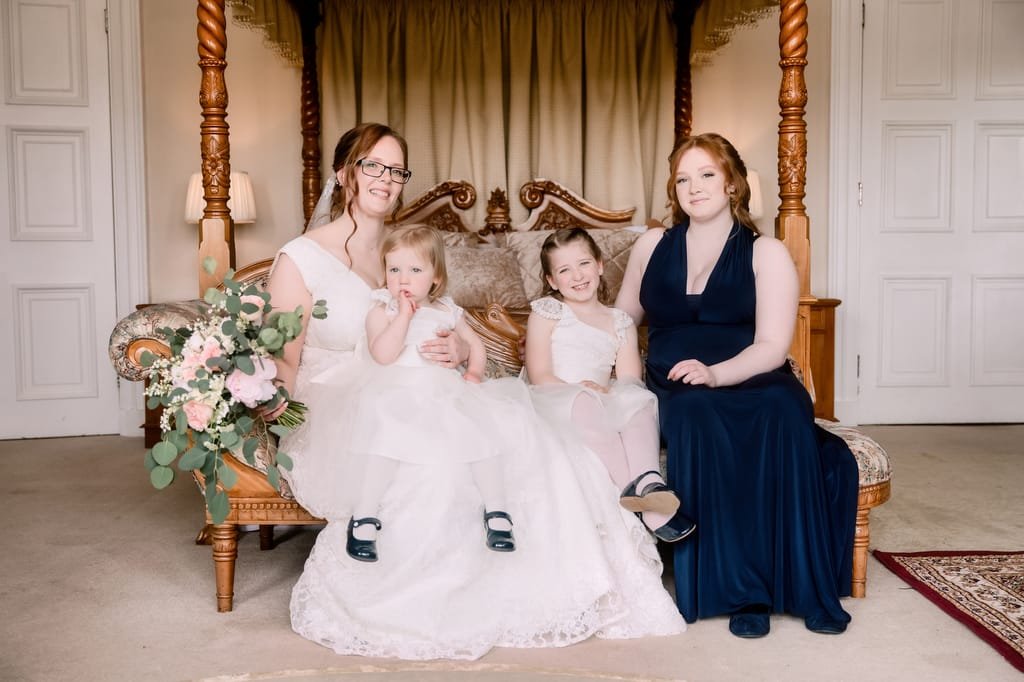 Four women of varying ages, dressed in formal attire for a wedding, sitting on an ornate sofa with floral decorations, in a well-lit room at Orchardleigh.