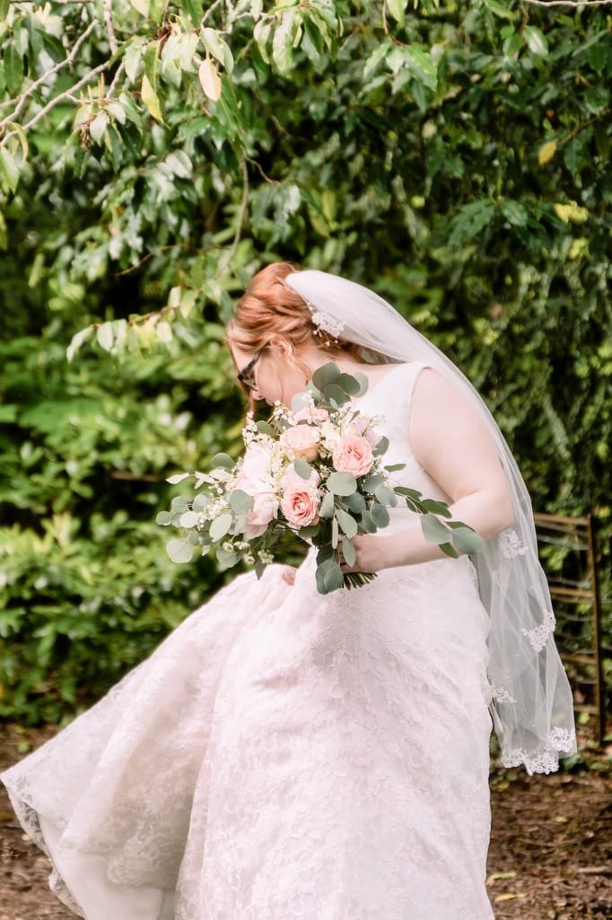 Bride in a white dress and veil smelling a bouquet of pink roses and greenery in a lush garden, captured by the wedding photographer at Orchardleigh.