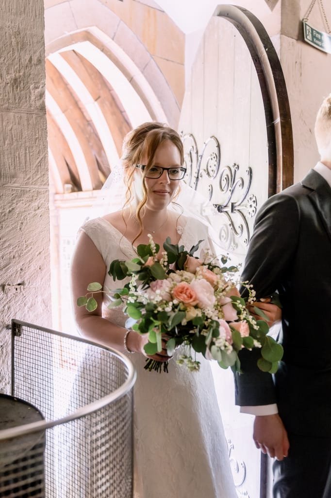 A bride holding a bouquet of flowers stands beside a groom in a sunlight-filled archway at Orchardleigh.