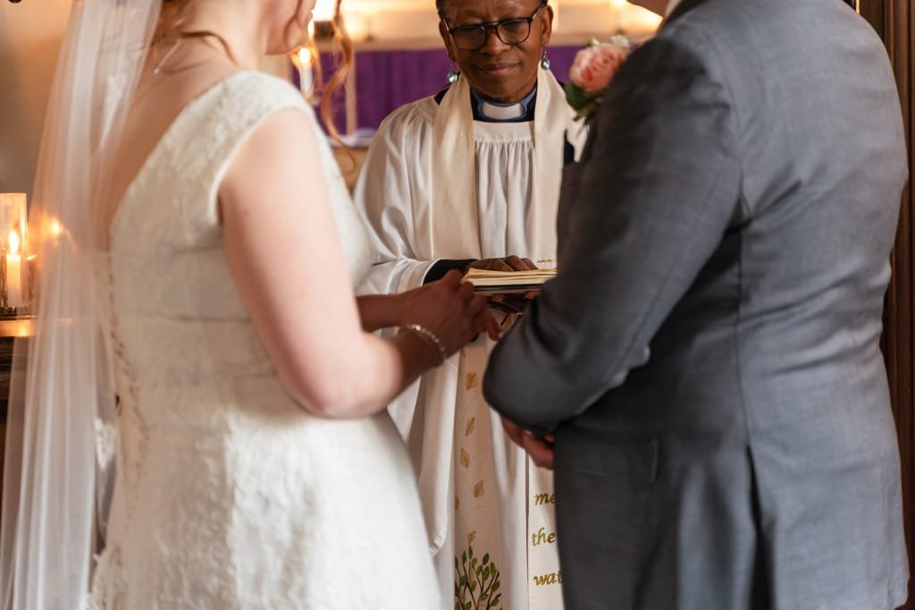 A priest officiating a wedding ceremony at Orchardleigh, holding a book as the bride and groom hold hands in front of him.