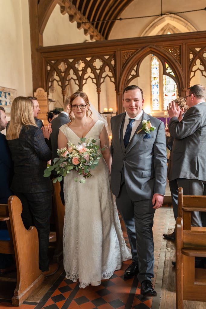 A bride and groom walk down the aisle of a church, holding hands and smiling, surrounded by applauding guests as a wedding photographer captures the moment.
