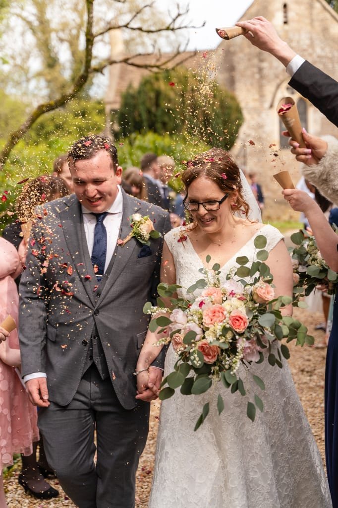 A bride and groom holding hands and smiling as they walk, surrounded by guests throwing confetti at them after a wedding ceremony captured by a skilled wedding photographer at Orchardleigh.