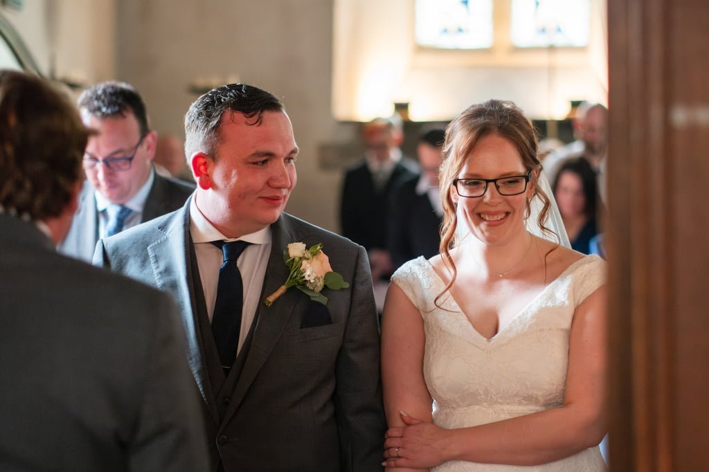 A bride and groom smiling and holding hands inside a church during their wedding ceremony, captured by a wedding photographer at Orchardleigh.