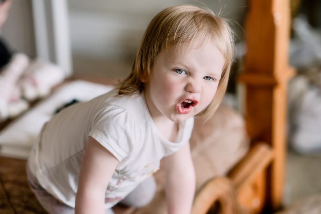 Toddler with blonde hair making a playful face, wearing a white T-shirt, sitting indoors by wooden furniture at Orchardleigh.
