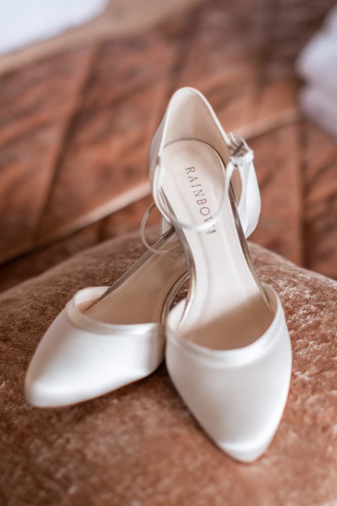 A pair of elegant white satin high-heeled shoes with ankle straps, ideal for a wedding photographer at Orchardleigh, displayed on a plush brown surface.