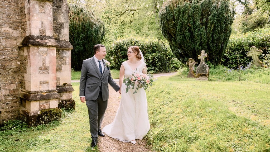 A couple in wedding attire walking hand in hand next to a stone building and lush greenery, captured by an Orchardleigh wedding photographer.