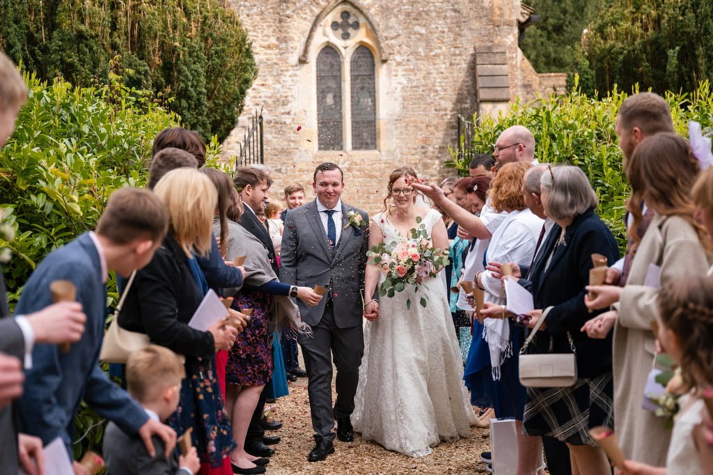 Newlywed couple walking down a path lined with guests outside a stone church, receiving congratulatory gestures from their wedding photographer at Orchardleigh.