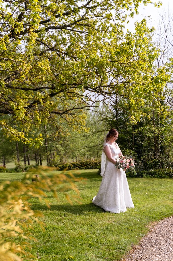 A bride in a white dress holding a bouquet stands on a grassy path under green trees in a sunny park, captured by a wedding photographer.