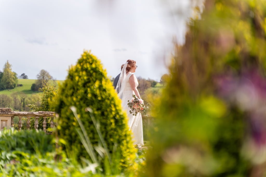 A bride holding a bouquet stands surrounded by greenery, gazing across the orchard on a sunny day.