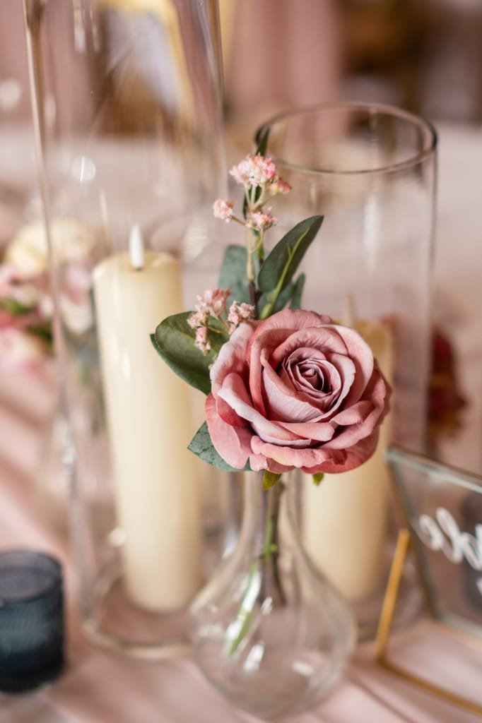 A delicate pink rose and small flowers arranged in a slender glass vase, captured by a wedding photographer at Orchardleigh, placed next to tall candles on a softly draped tablecloth.