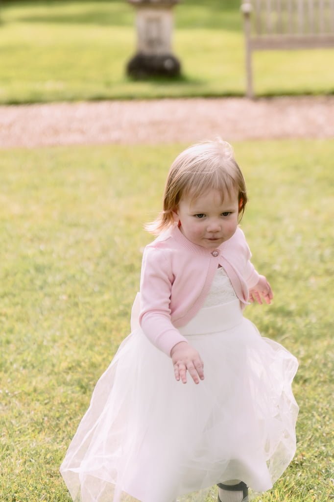 A toddler girl in a white dress and pink cardigan walks on a grassy area with a soft focus background featuring an orchardleigh garden and gate.