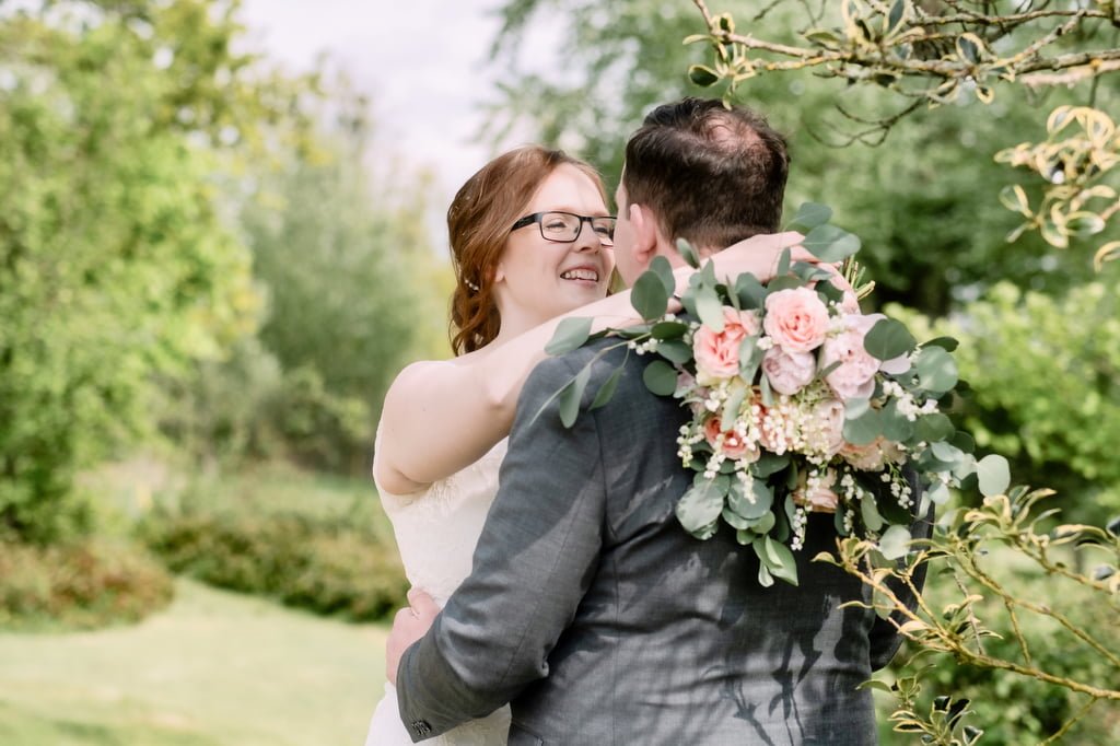 Bride and groom smiling at each other, holding a bouquet, outdoor setting with greenery in the background captured by the wedding photographer at Orchardleigh.