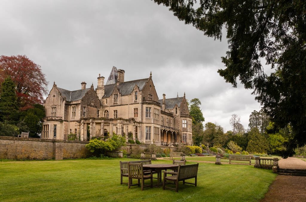 A large, historic stone mansion with multiple chimneys, surrounded by lush gardens and wooden patio furniture in the foreground, ideal for a wedding photographer at Orchardleigh.