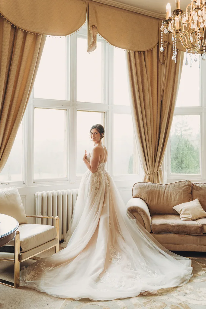 Orchardleigh183 A bride in an elegant white dress with a long train stands by a window in Orchardleigh House, a well-appointed room.